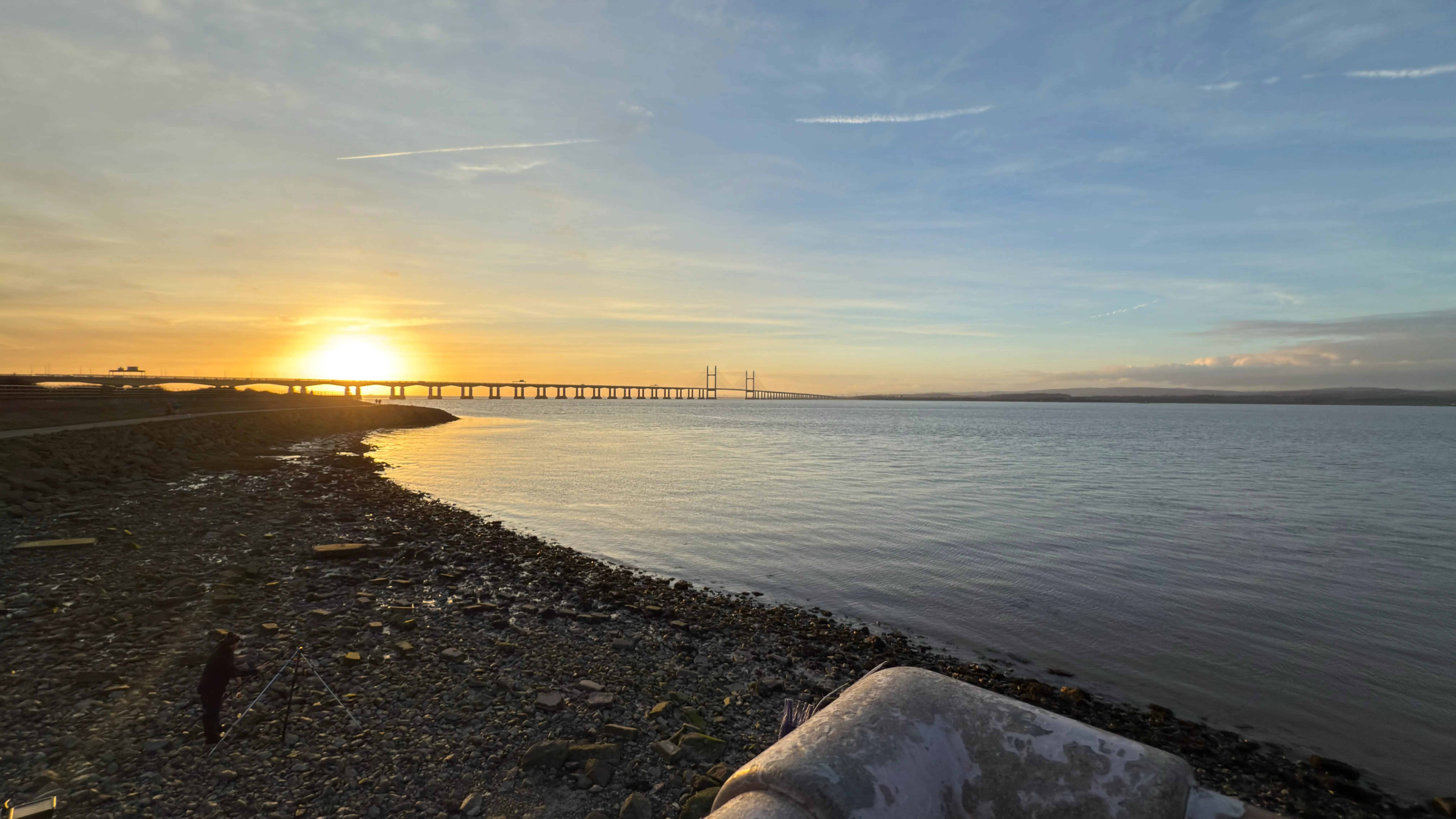 A wide angle photograph of the Prince of Wales Bridge (formerly, the Second Severn Crossing) at around sunset. Most of the bridge's length is visible in silhouette above the Severn Estuary, with a rocky shore in the bottom-left sixth of the photo.