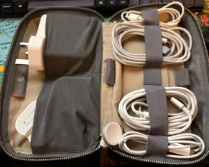 Top down photograph of a rectangular, tan-coloured bag that has been laid open. In two pockets on the left side are a large laptop 'wall wart' and two folding wall socket adaptors. On the right are four cables of varying lenghts and types wound into ovals and held together by elasticated straps.