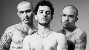 Black and white photo of three topless men. The leftmost model has a closely shaved head and a mustache, nose and ear piercings, and a number of tattoos on his face and torso. The central model has medium length curly hair and a beard, ear piercings and a couple of tattos. The right model has a shaved head, a goatee, a number of torso tattoos and some elaborate over-ear hoop piercings.