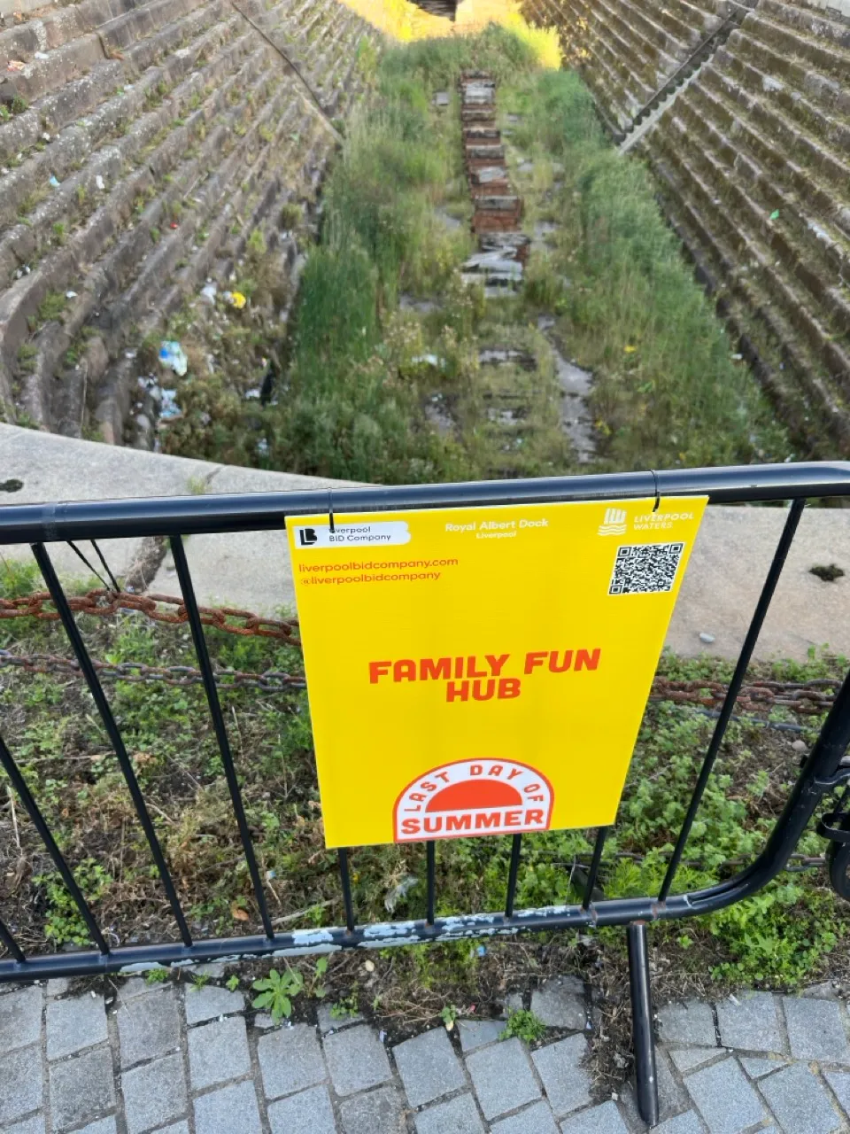 Looking down into a deep boat dry dock made of roughly hewn rock, overgrown with grass and filled with trash. A sign attached to a fence outside it reads 'Family Fun Hub'.