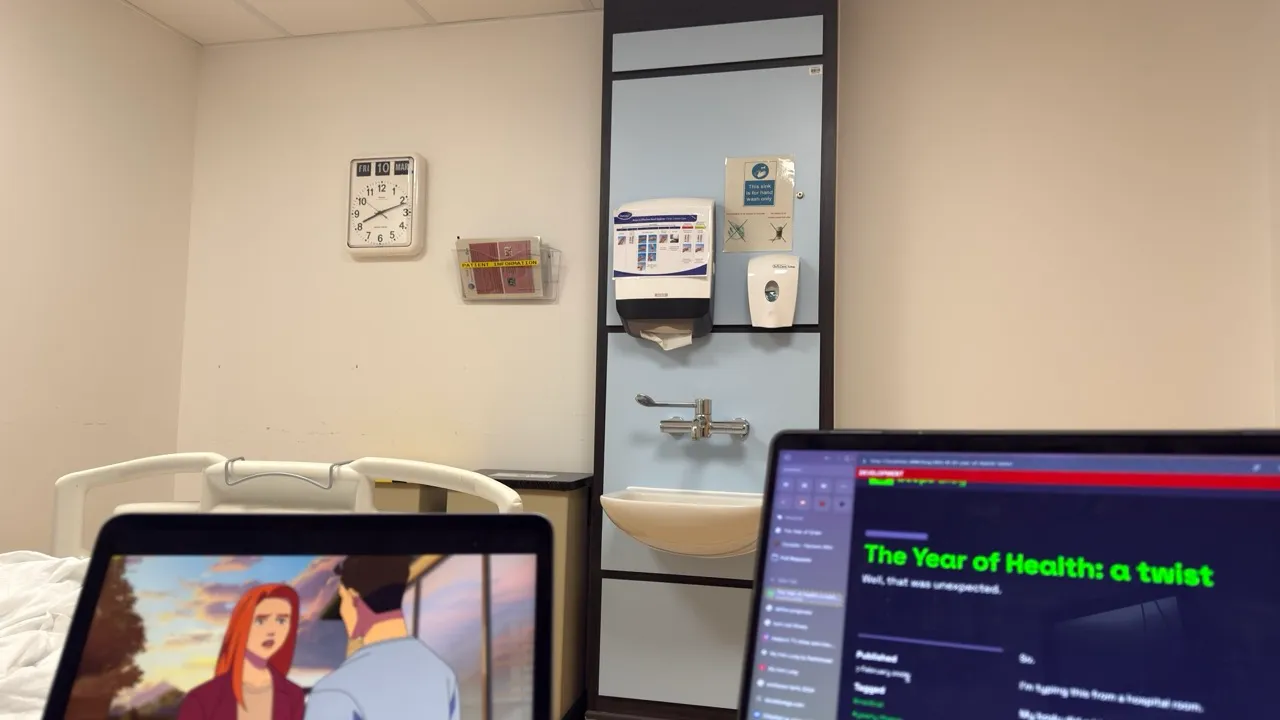 Photograph of my hospital room. In the foreground is an iPad showing an episode of Invincible and a laptop with this blog post open. In the background is a clock that shows the date March 10th for some reason.