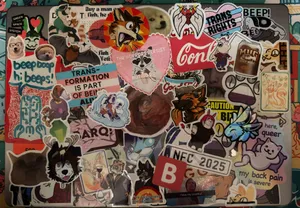 Top down photograph of a laptop lid which has been covered nearly edge to edge with an overlapping patchwork of stickers. Includes numerous stickers of people's fursonas, various animals, trans and agender pride flags, and humorous motivational phrases.